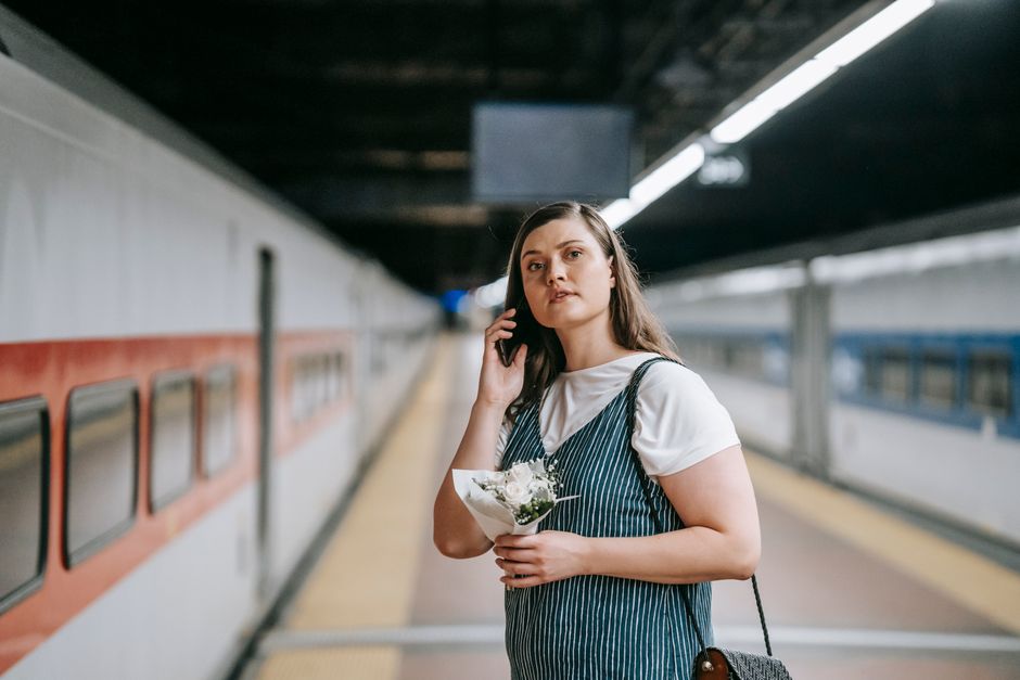 A young woman with long, dark brown hair and fair skin stands on a train station platform, holding a small bouquet of white flowers wrapped in white paper with green foliage accents in her left hand. 