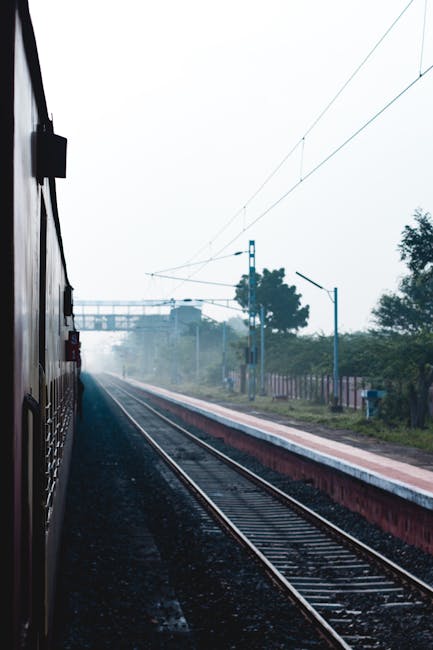 The image depicts an outdoor train station platform adjacent to railway tracks, with a train visible on the left side partially out of frame. The scene is overcast with a soft, diffuse light creating 