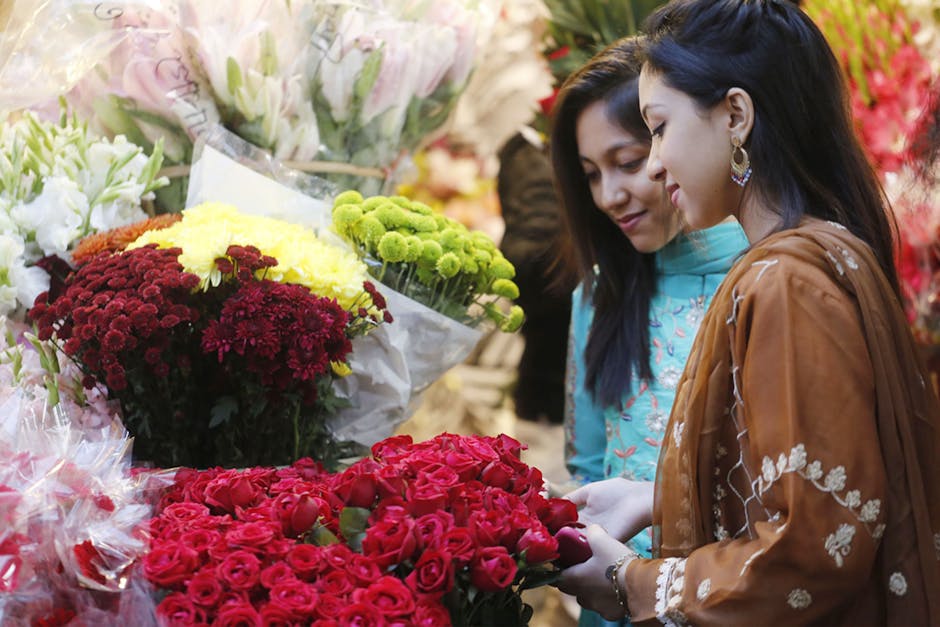 Two women examining a vibrant bouquet of red roses in a florist shop. The arrangement features fresh, tightly packed roses with velvety petals, displayed on a table surrounded by various floral bouque