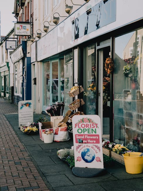 Exterior view of a flower shop on Main Road Romford with large glass windows displaying floral arrangements inside. Outside the shop, there are buckets and baskets filled with freshly cut flowers, inc