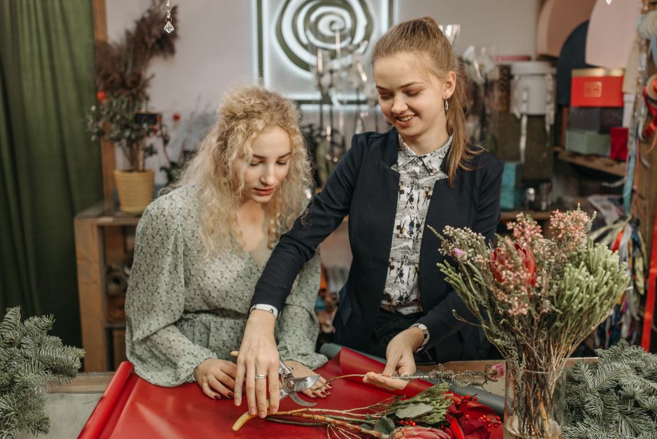 A floral arrangement workshop showing two women inside a florist shop. The woman on the right, wearing a dark blazer over a patterned blouse, is holding a pair of scissors and is arranging a bouquet o