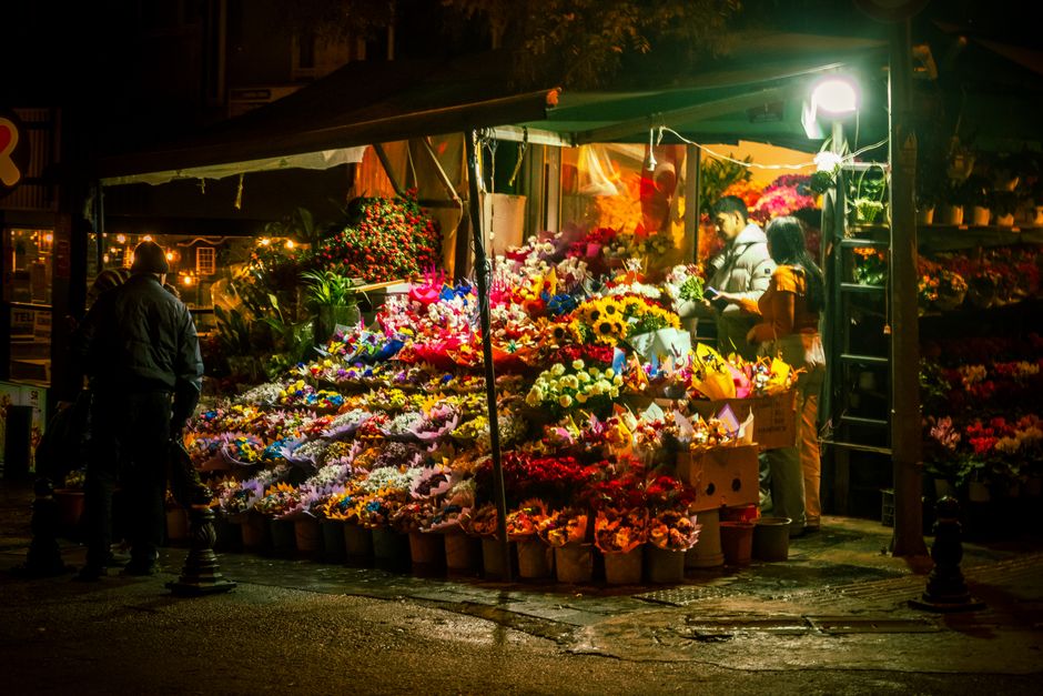 A flower stall at night illuminated by a bright white light, displaying a vibrant array of fresh flowers in various colors, including yellow sunflowers, pink, white, and purple blooms, arranged in bou